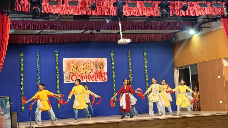 Dancers in yellow costumes and red scarves in a line across a stage with a blue backdrop and a Dance Competition banner