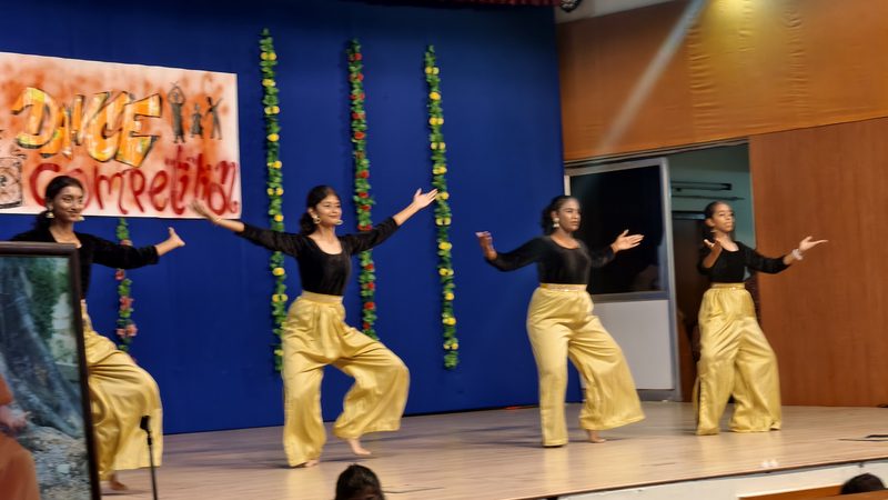 Four dancers in black tops and gold trousers striking a pose on a stage with a blue backdrop