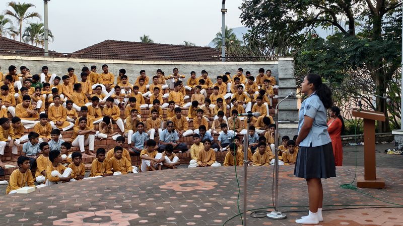 A student in school uniform reciting at a microphone on a tiled terrace, tiers of pupils in mustard kurtas seated behind