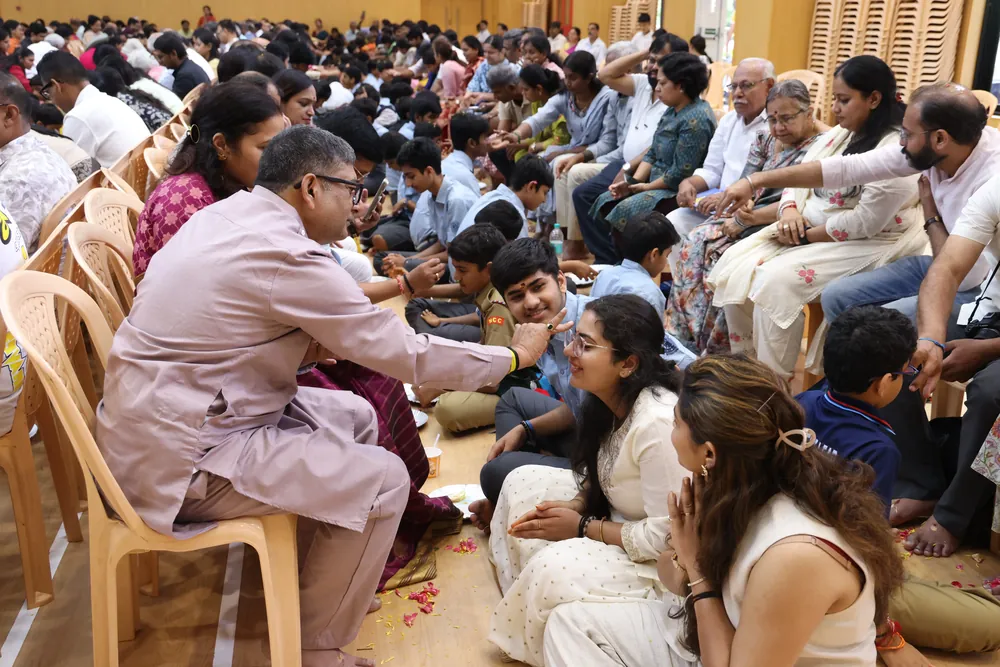 Parents seated on chairs in a crowded hall, with children sitting on the floor in front of them and flower petals scattered around; some parents reach toward their children’s faces