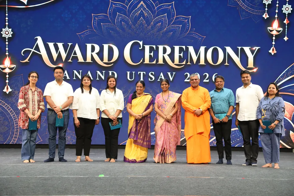 Ten people standing in a row on a stage under a blue backdrop that reads Award Ceremony, Anand Utsav 2026, a swami in saffron among them