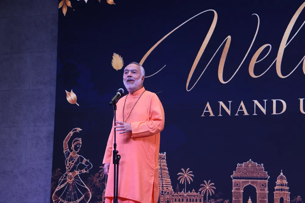 A swami in saffron speaking into a microphone on stage, in front of a backdrop that begins with the word Welcome and reads Anand Utsav