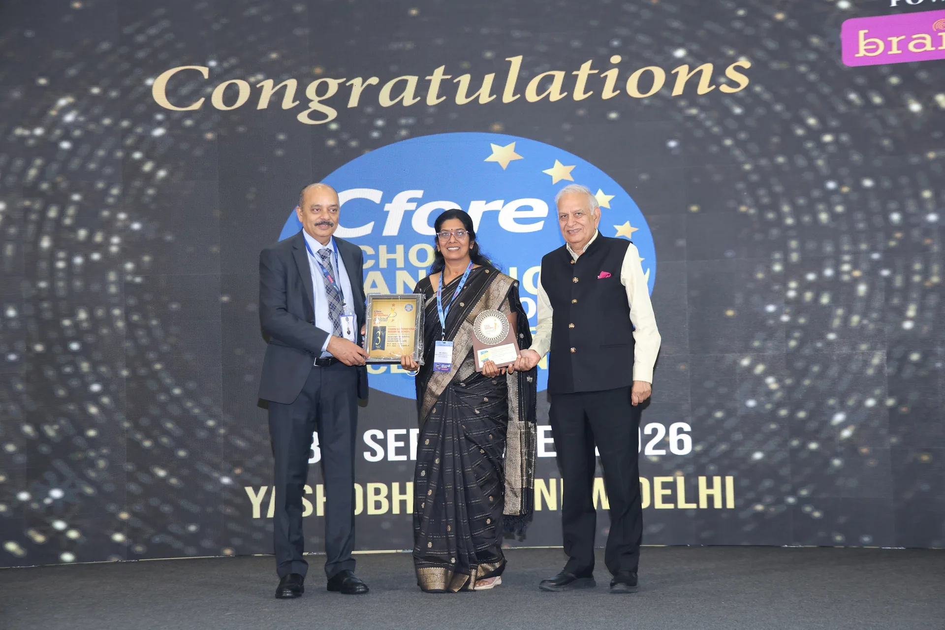 A woman in a sari holding a trophy on the stage of the Cfore School Rankings 2026 event, between two men