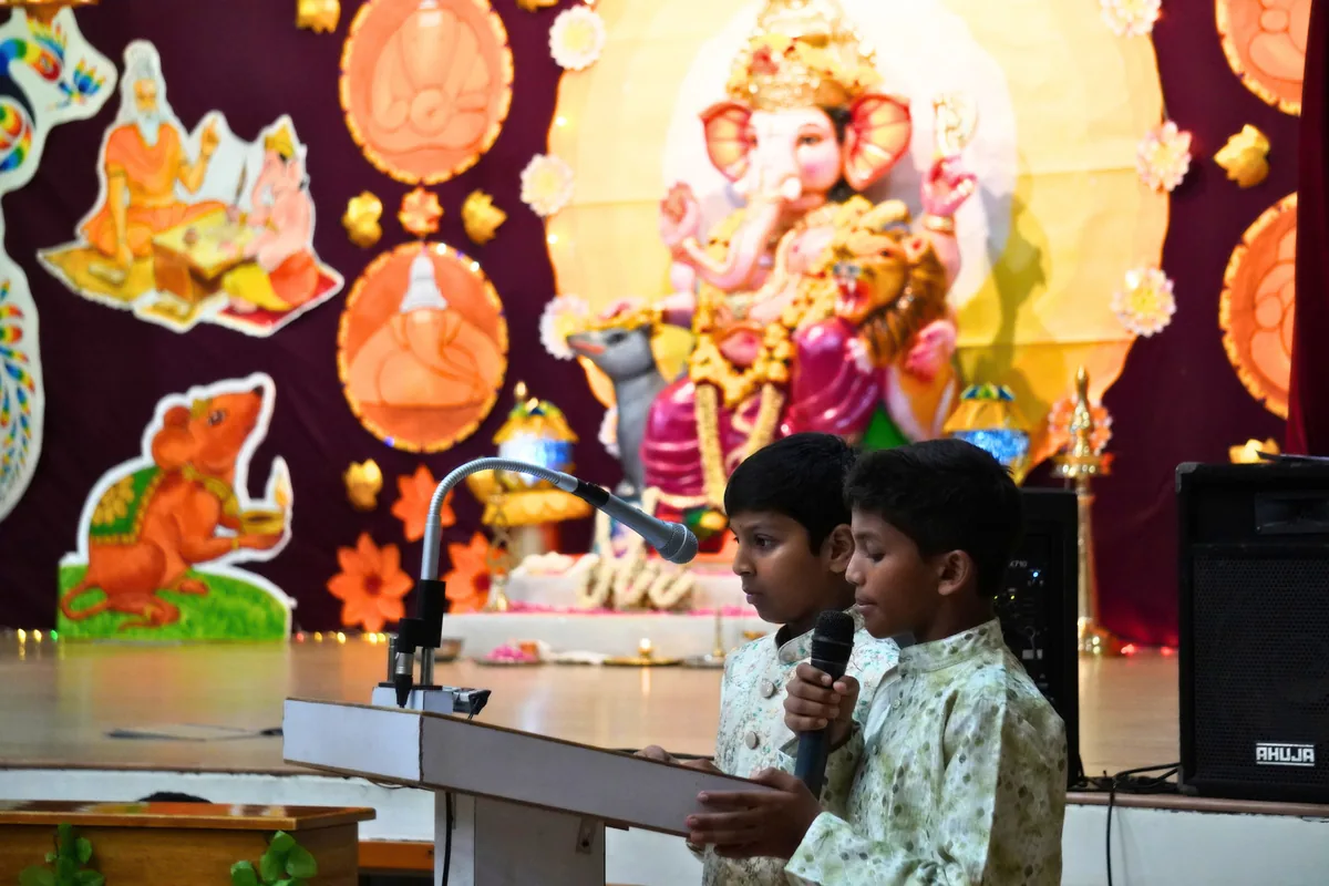 Two boys at a lectern before a decorated Ganesh backdrop