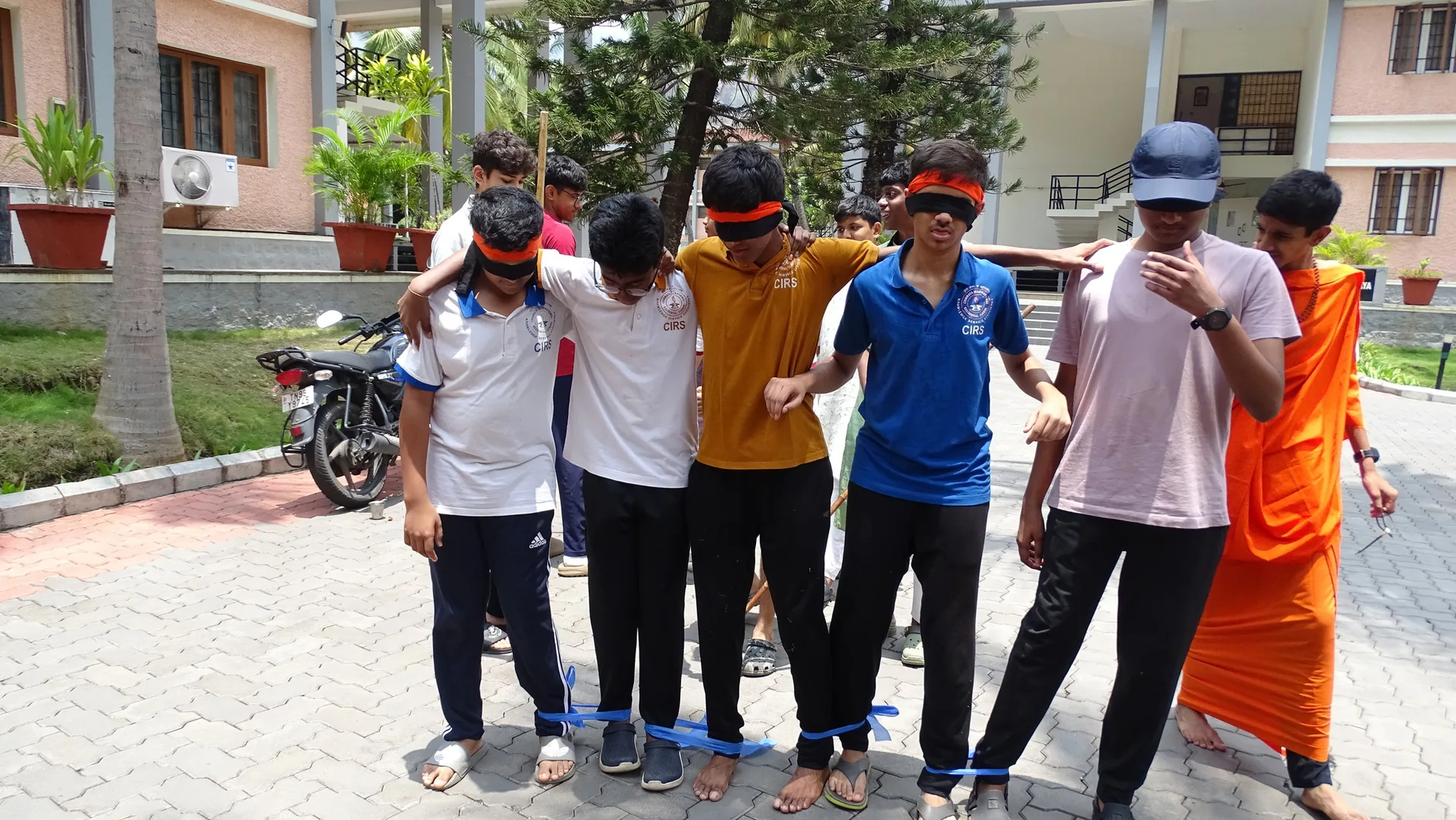 Students standing in a row in a courtyard wearing coloured bands, with a person in saffron beside them