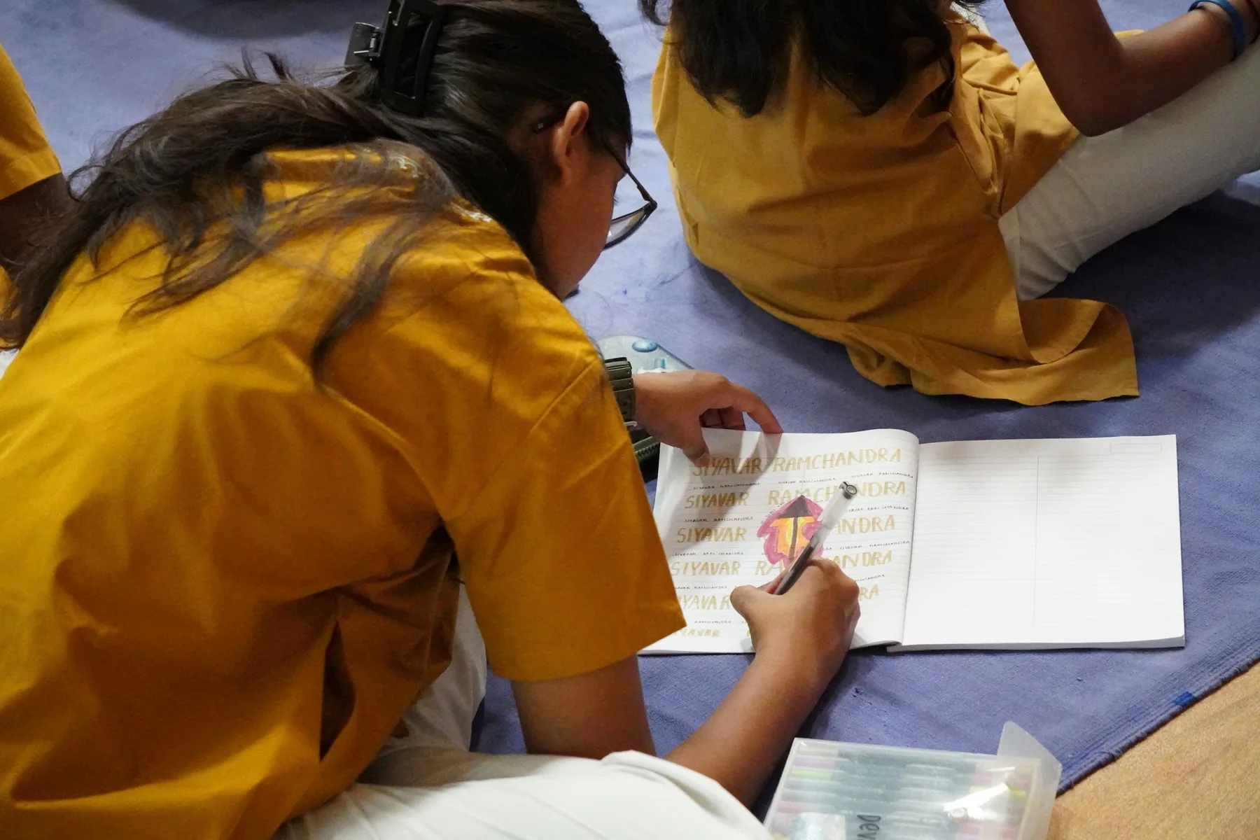A student in a yellow kurta writing in a notebook, seated on the floor