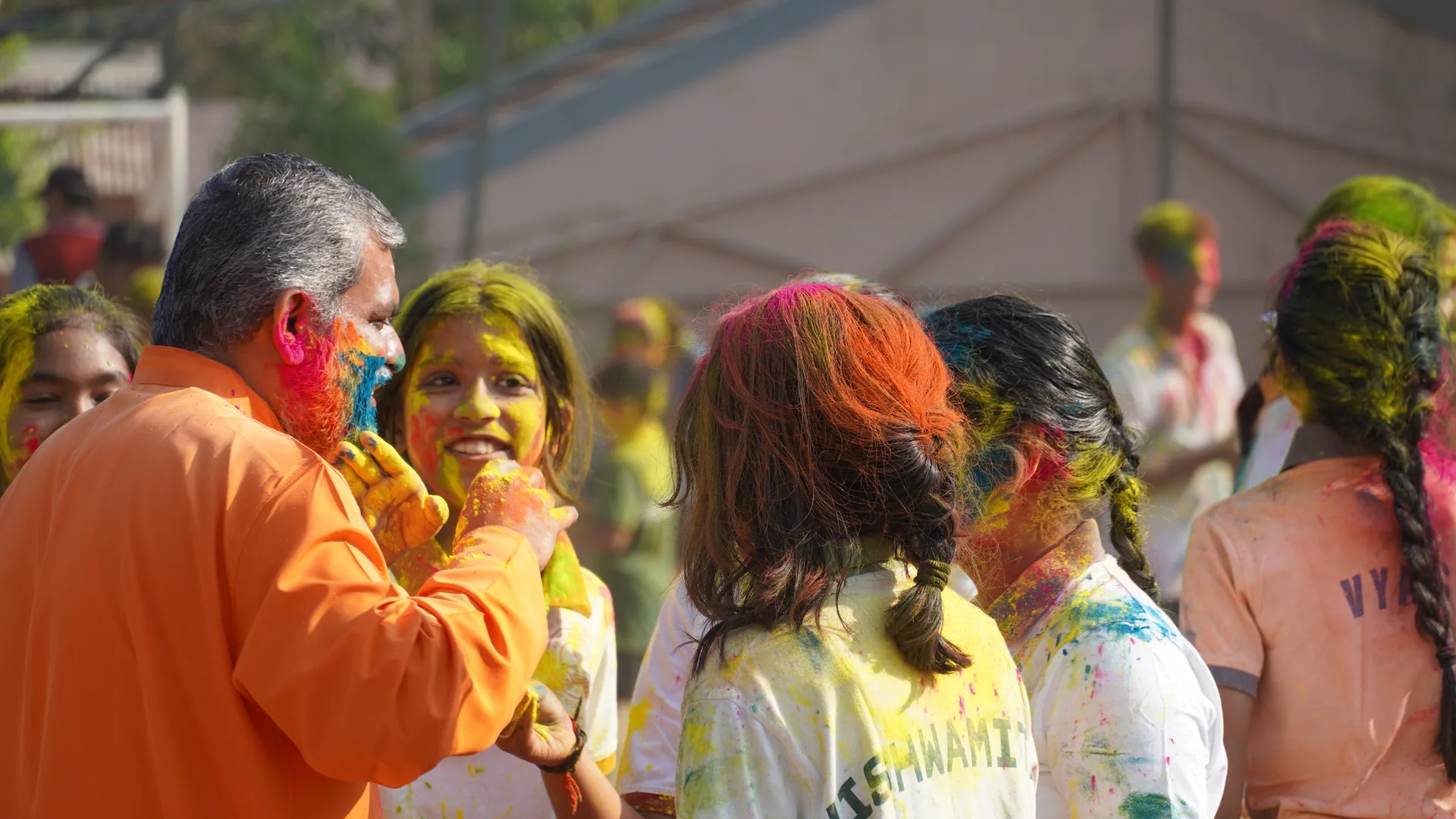 A man in an orange kurta smearing colour on a smiling student among others with coloured faces at Holi