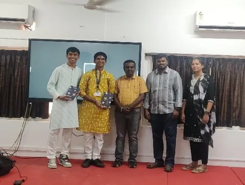 A student in a white kurta and another in a yellow kurta with three adults before a screen