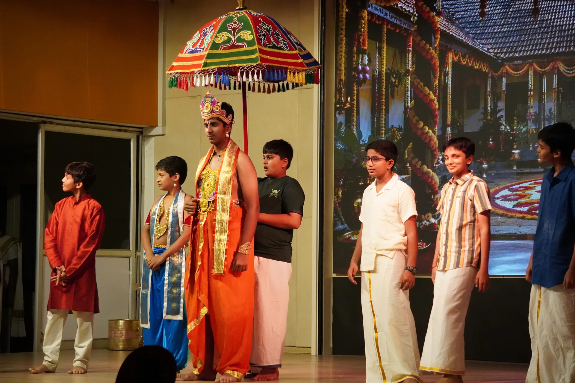 Students on a stage in a skit, one standing beneath a colourful ceremonial umbrella
