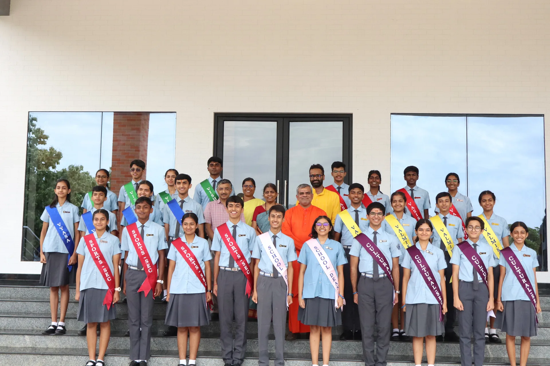 The Student Council 2026–27 standing in rows on the steps of a school building, in sashes over school uniform, with a swami in saffron among them