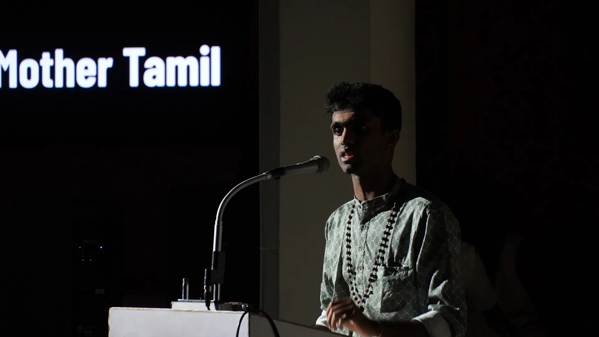 A student speaking at a microphone beneath a sign that reads Mother Tamil