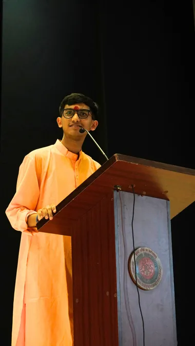 A student in a saffron kurta speaking at a lectern