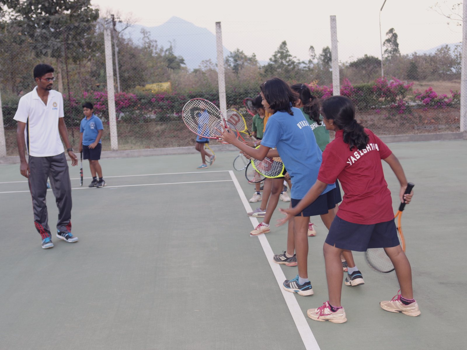 A coach watches a row of CIRS students practise their forehand swing with tennis racquets on a green hard court