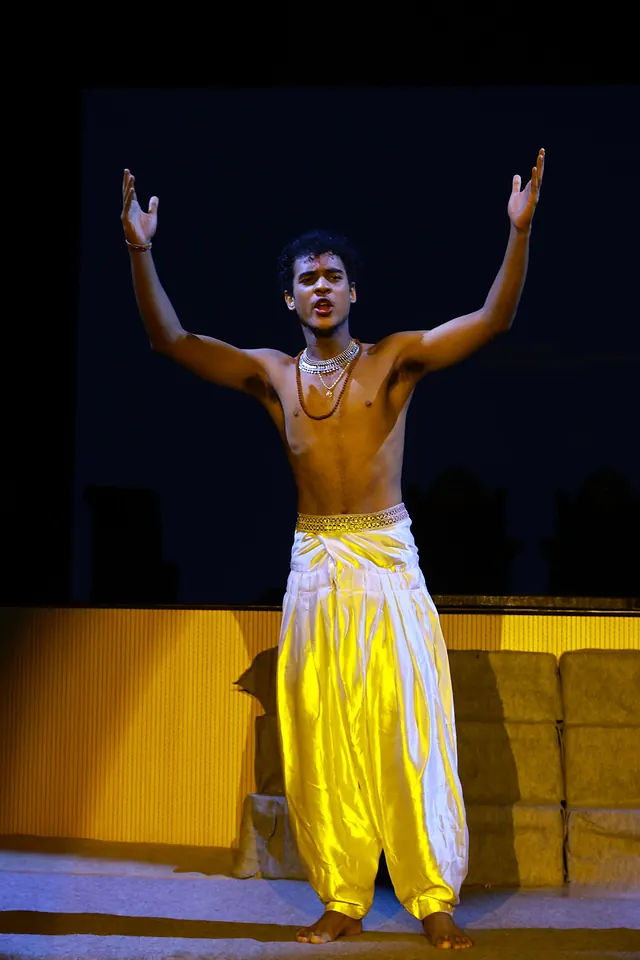 A student in a yellow dhoti alone in a pool of light on a dark stage, both arms raised