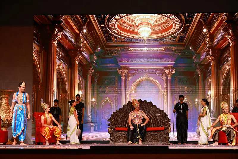 A palace hall filling the stage: a king on a carved throne, courtiers in bright costume and guards in black around him