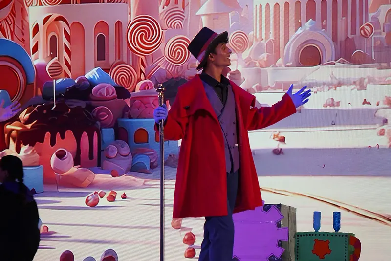 An actor in a red coat and top hat, arm out, before a pink candy-land backdrop