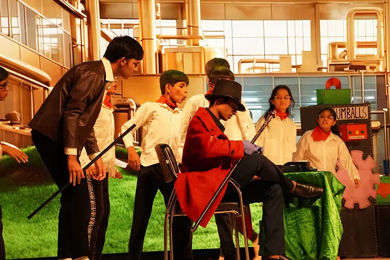 Actors in a factory set, one in a red coat seated with a cane