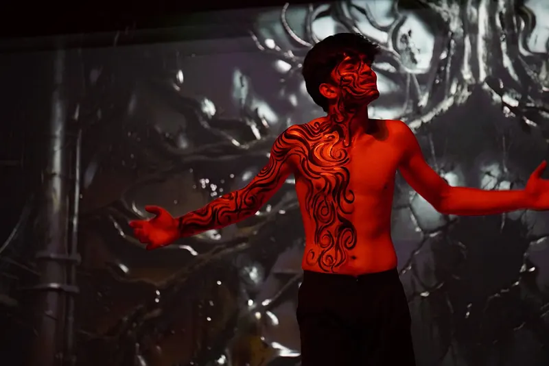 An actor in red swirling body paint, arms spread, against a dark backdrop