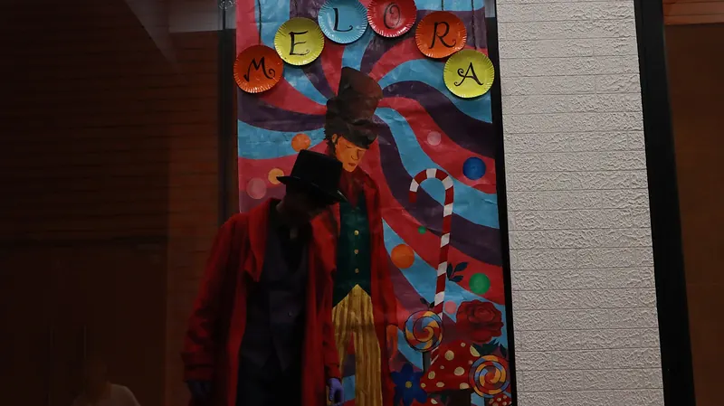 A figure in a red coat and top hat before a painted poster reading Melora