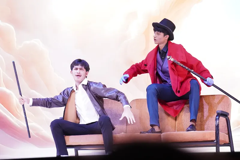 Two actors on a sofa before a white-and-peach backdrop, one in a red coat holding a cane