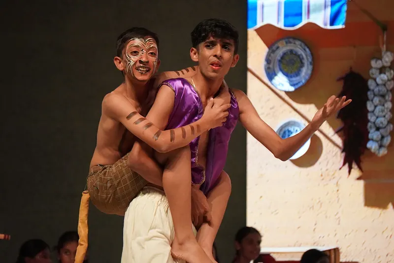 One actor climbing onto another's back beside a wall of hanging plates