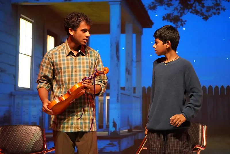 An actor holding a violin and a boy on a porch set at night
